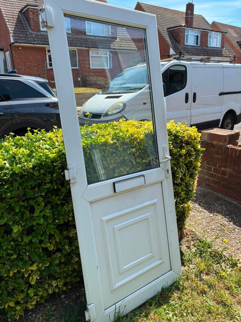 Full view of a removed white UPVC residential door resting against a garden hedge prior to new hinge fitting
